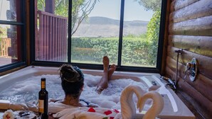 Une pièce dotée d’une grande fenêtre, d’un décor intérieur en bois, et d’une baignoire remplie de pétales de roses et décorée de motifs en forme de cœur.