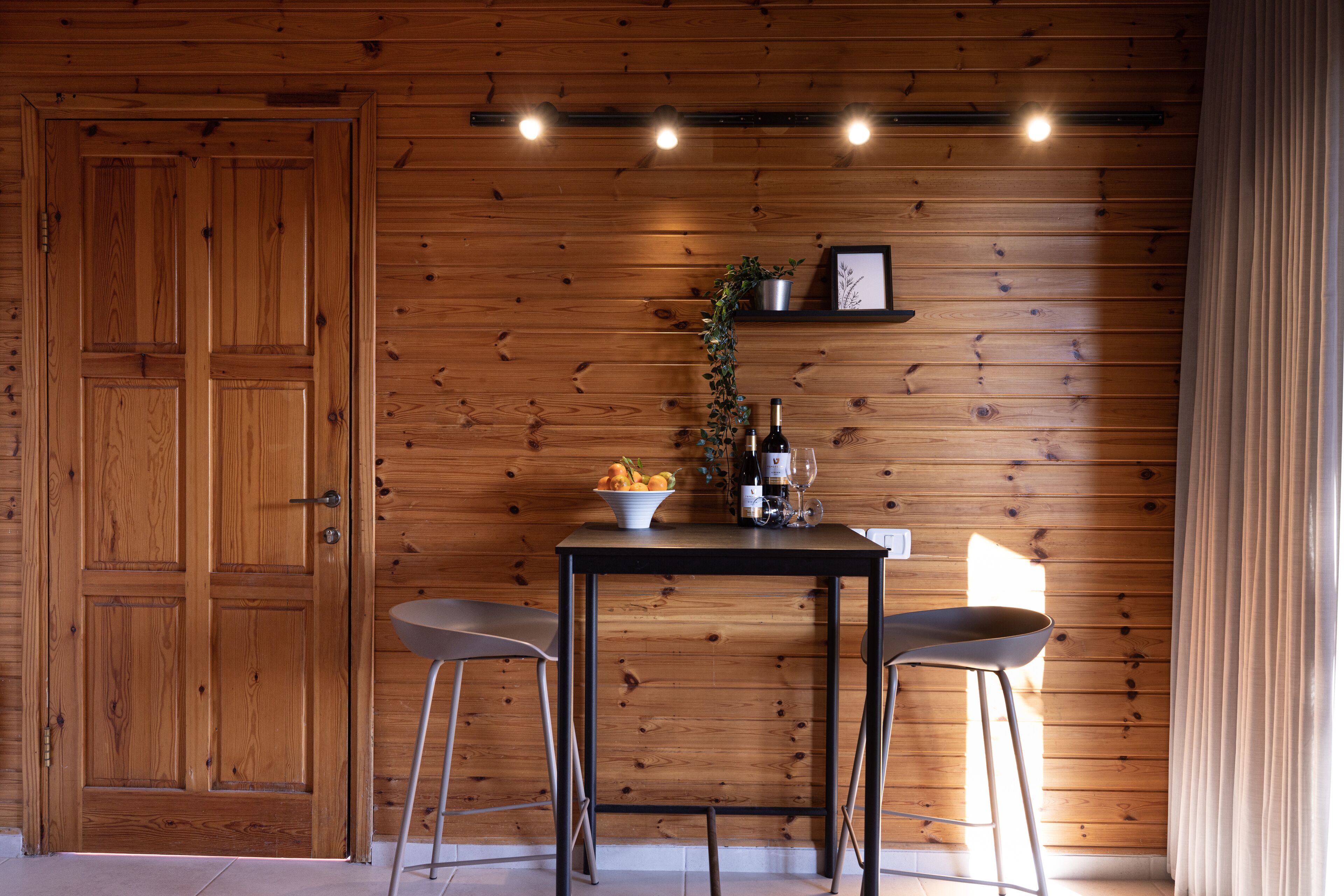 A wooden-paneled room with a table, two chairs, a plant, and a shelf with bottles and a picture frame.
