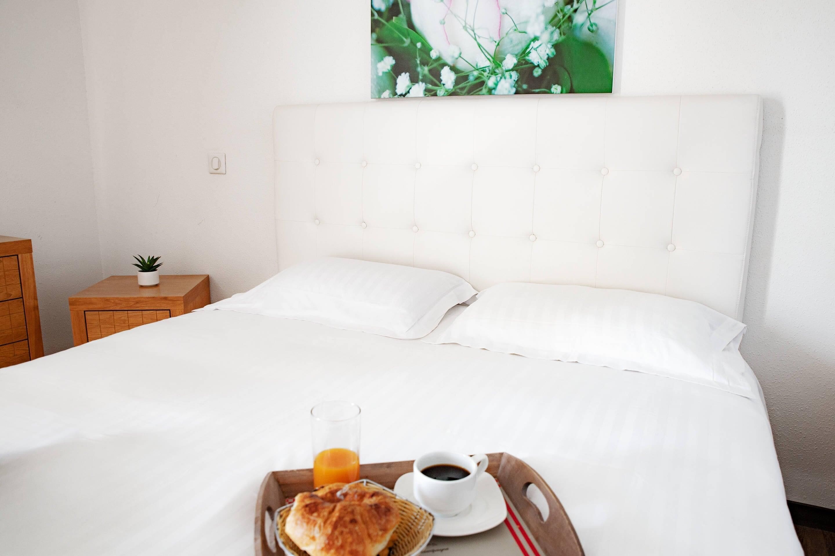 En pænt redt seng med hvide sengetæger, et bakke med morgenmadsprodukter, heriblandt en croissant, et glas appelsinjuice og en kop kaffe.