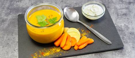 Un bol de soupe jaune, agrémenté d’une garniture, une cuillère et un petit bol de sauce blanche, le tout présenté sur une planche d’ardoise avec des bâtonnets de carottes.