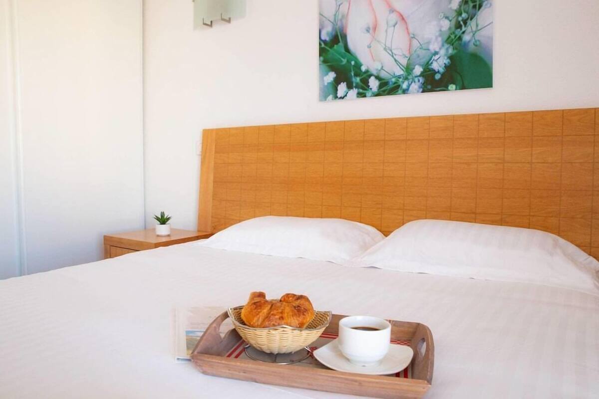 Una cama bien hecha con sábanas blancas, una bandeja con cruasanes y una taza de café, y una pequeña planta en una mesita de noche.
