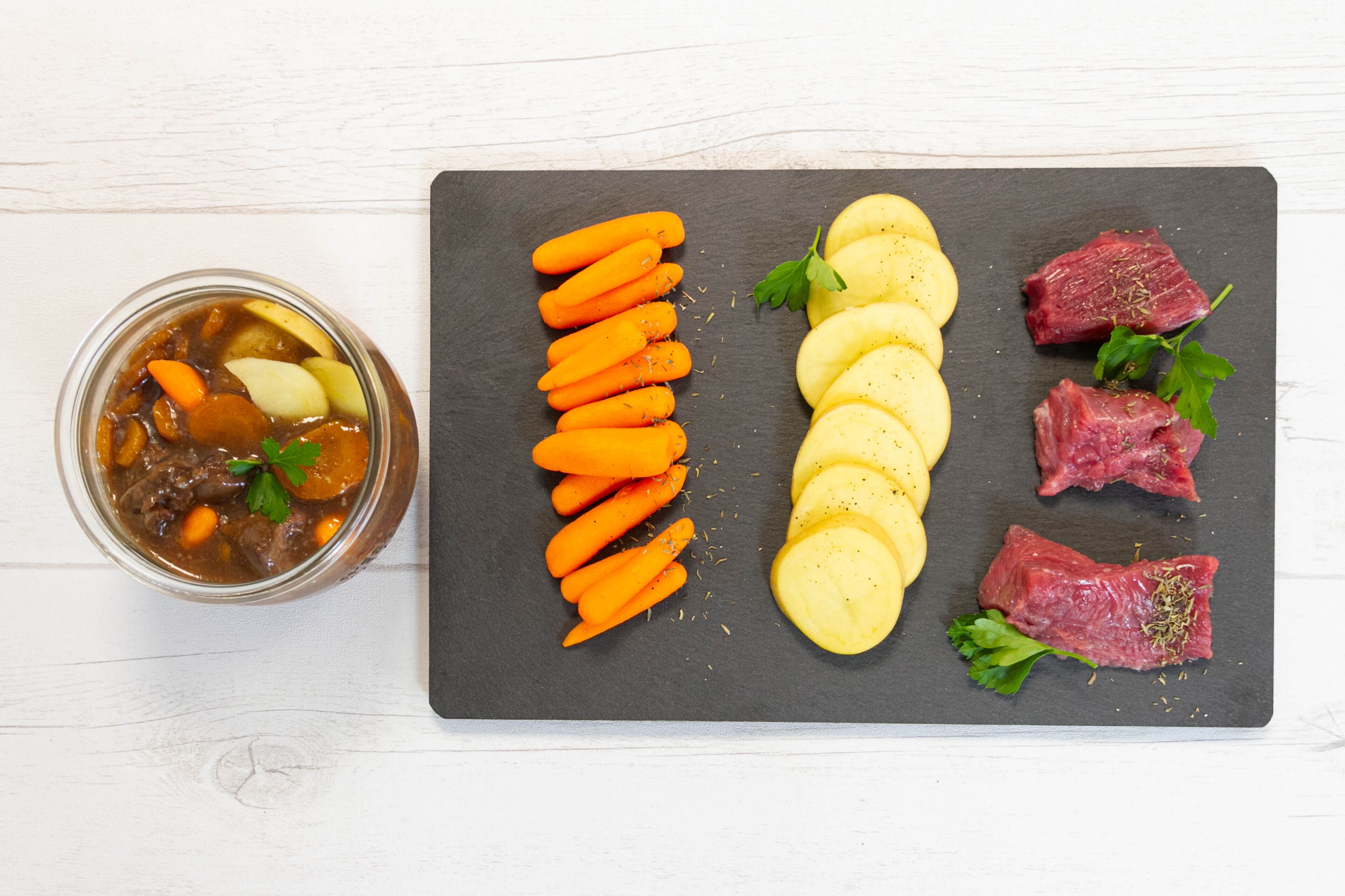 A meal with sliced carrots, potatoes, and beef on a slate board, and a jar of stew with vegetables on the side.