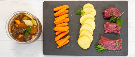 A meal with sliced carrots, potatoes, and beef on a slate board, and a jar of stew with vegetables on the side.