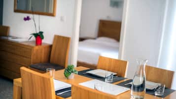 A hotel room with a wooden dining table set for two, a bed in the background, and a small potted plant on the table.