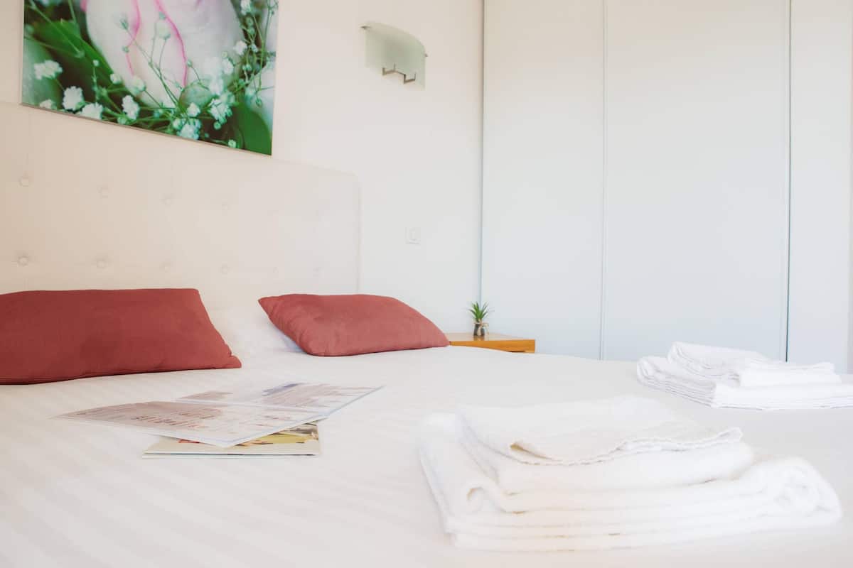 Una cama bien hecha con almohadas rojas, una mesita de noche con una planta y una pila de toallas blancas.