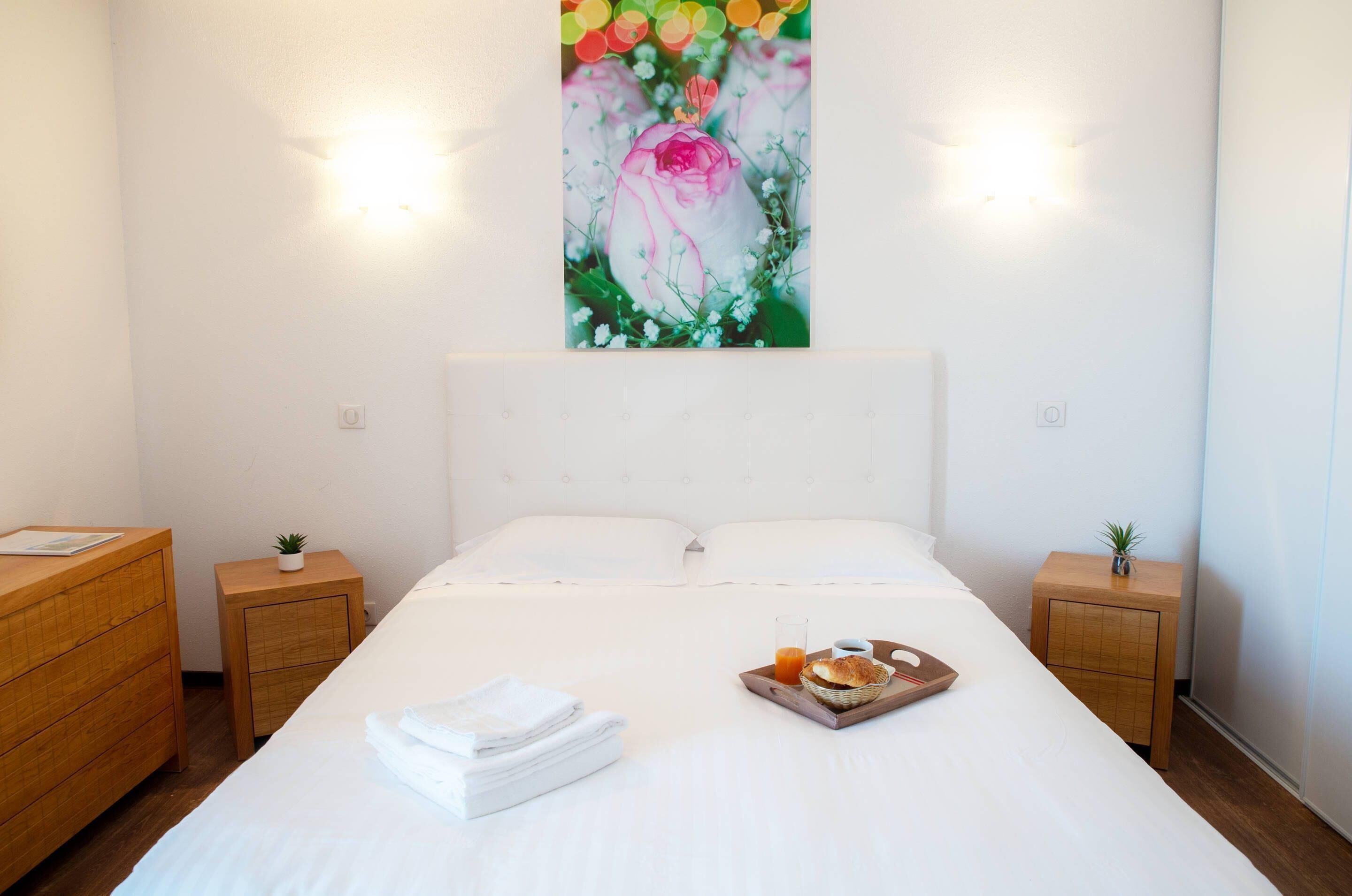 Ein Hotelzimmer mit einem großen Bett, zwei Nachttischen aus Holz, einem Wandbild und einer Ablage mit Frühstücksgegenständen.