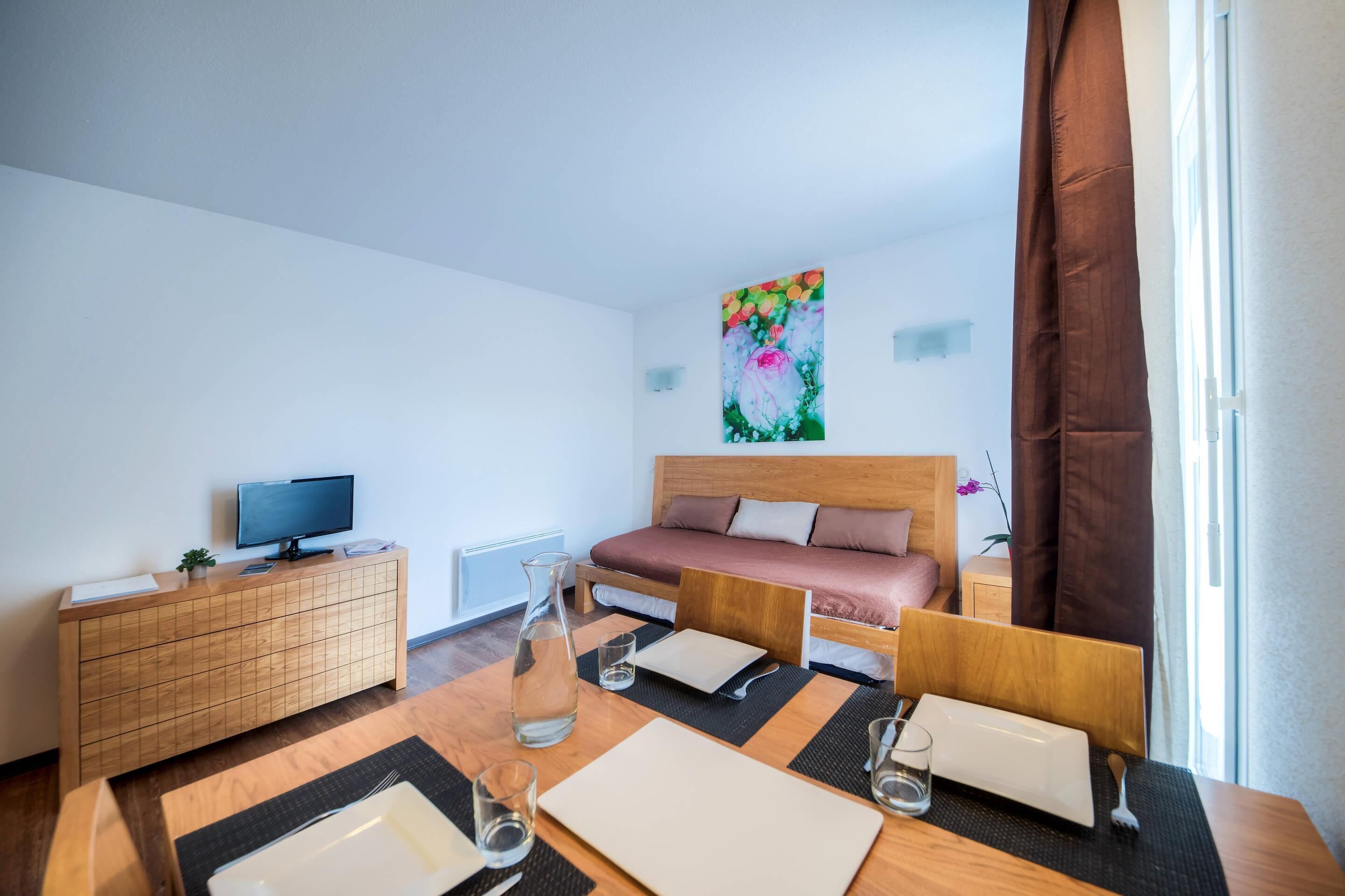 Moderni hotellihuone, jossa on puinen sänky, puinen tv-taso, ruokapöytä maljakko täynnä kukkia ja seinällä taulu.