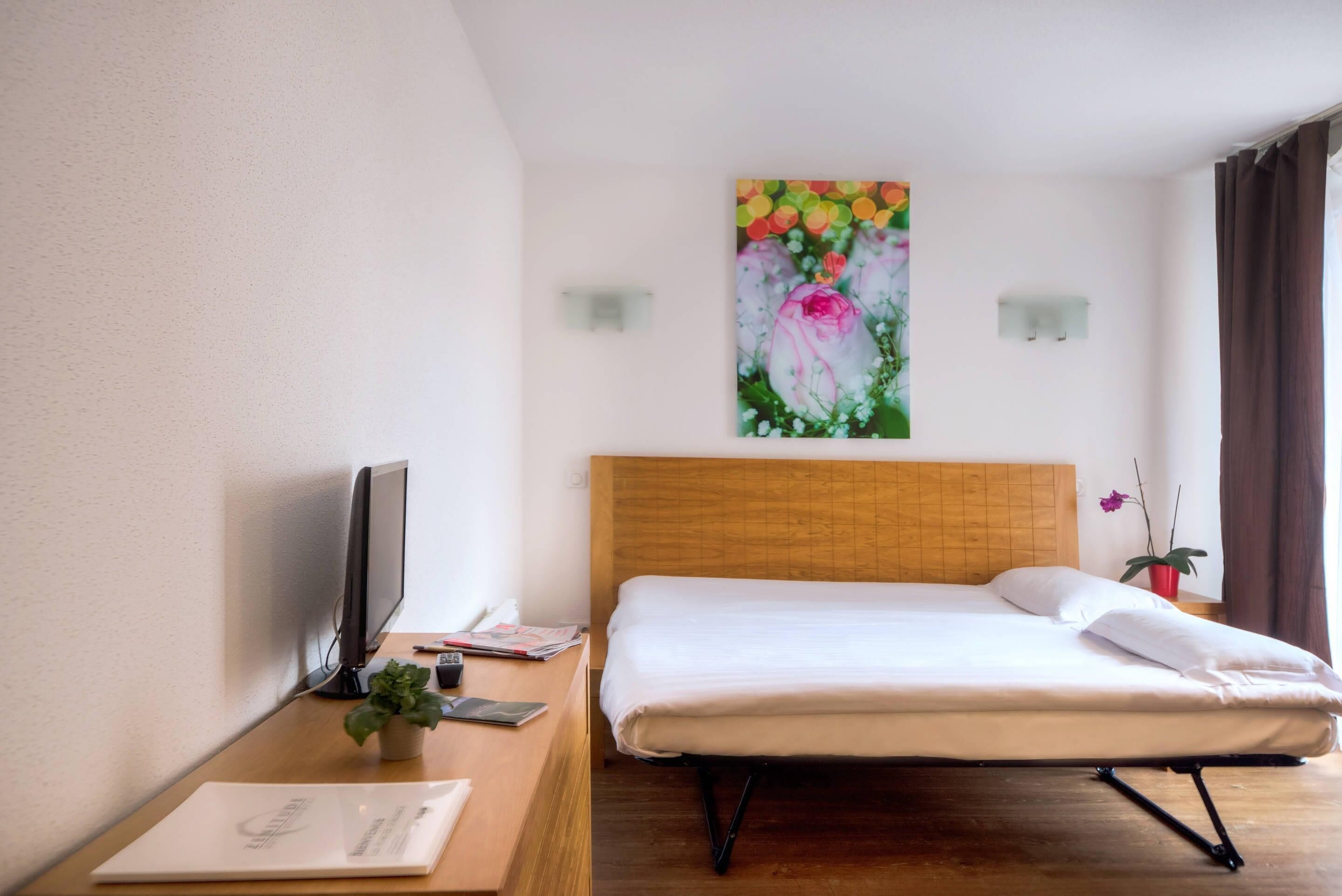 A hotel room with a wooden bed, a desk with a computer, a TV, and a painting of flowers on the wall.