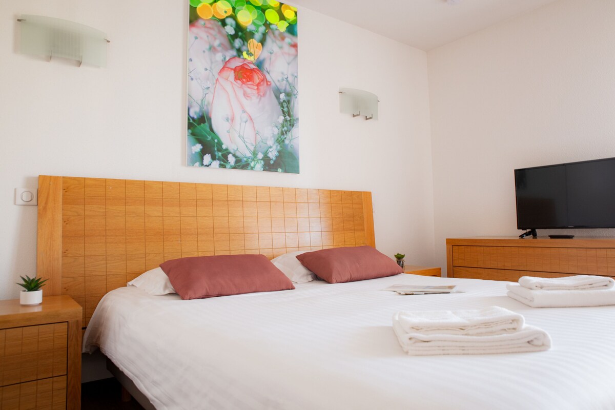 Habitació d'hotel amb un panell de fusta al capçal, un televisor de pantalla plana, un quadre amb flors, una petita planta i tovalloles doblegades amb cura.