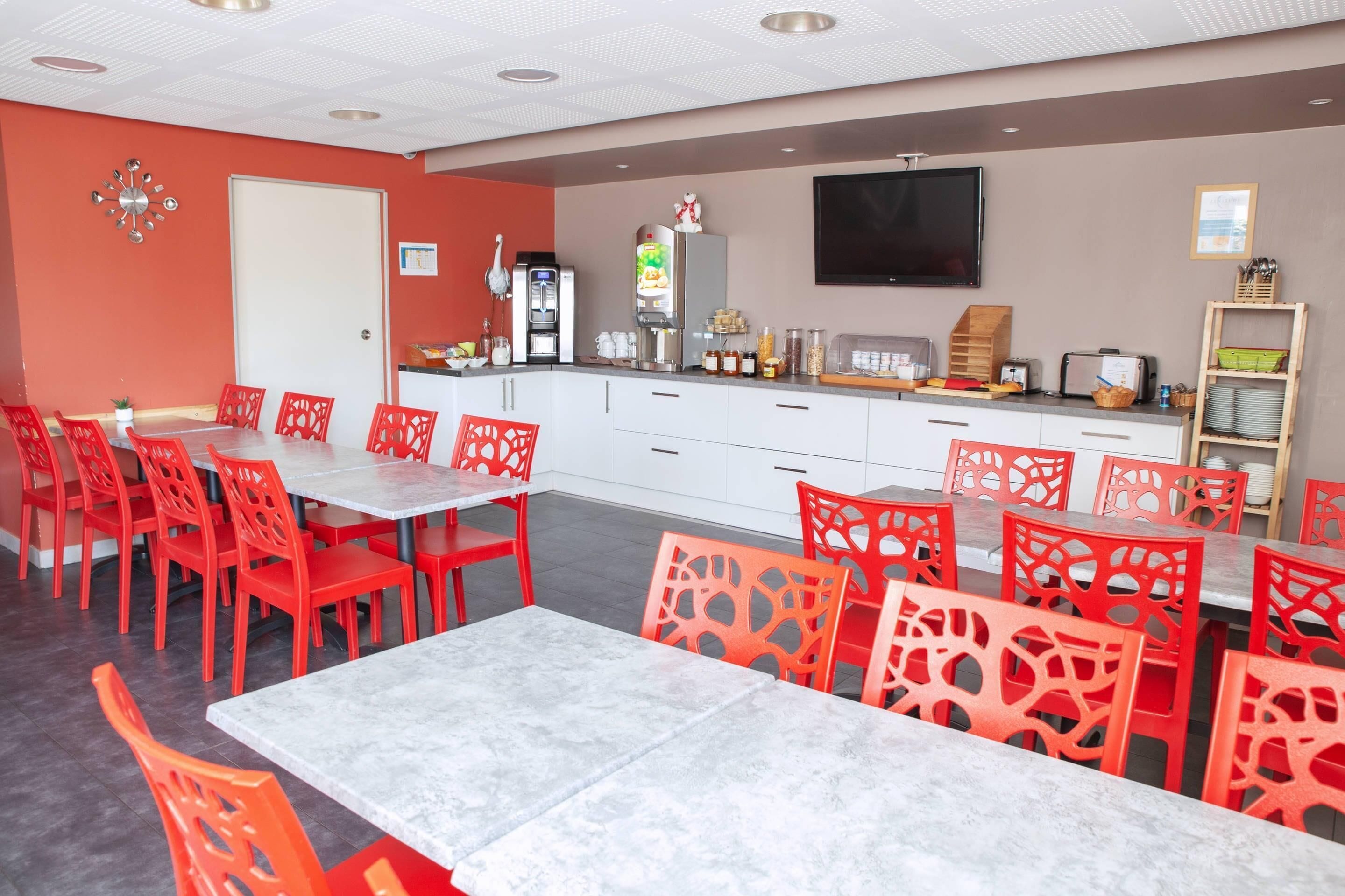 A dining area with red chairs and tables, a kitchen counter with appliances, and a television mounted on the wall.