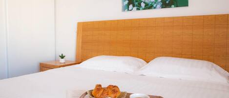 A neatly made bed with white linens, a wooden headboard, a tray with croissants and a cup of coffee, and a small plant on a bedside table.