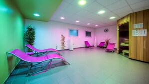 Una habitación con una pared verde, un panel de madera y un techo con iluminación empotrada. Hay dos sillones de masaje rosa, una planta en maceta y un reloj de pared.