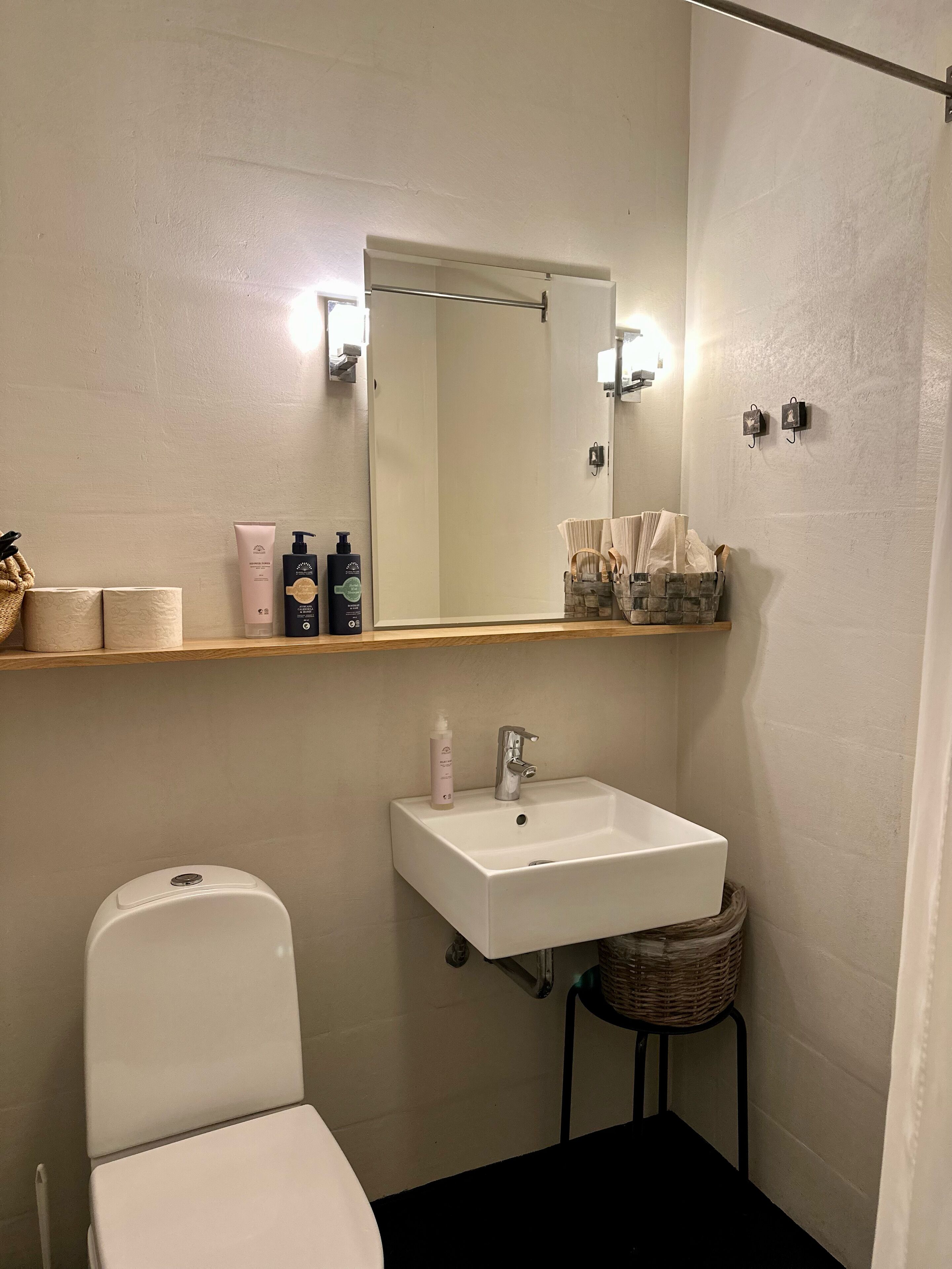 Une salle de bain avec des toilettes blanches, un lavabo blanc et un miroir au-dessus du lavabo. On trouve des articles de toilette sur une étagère et un panier sur le sol.