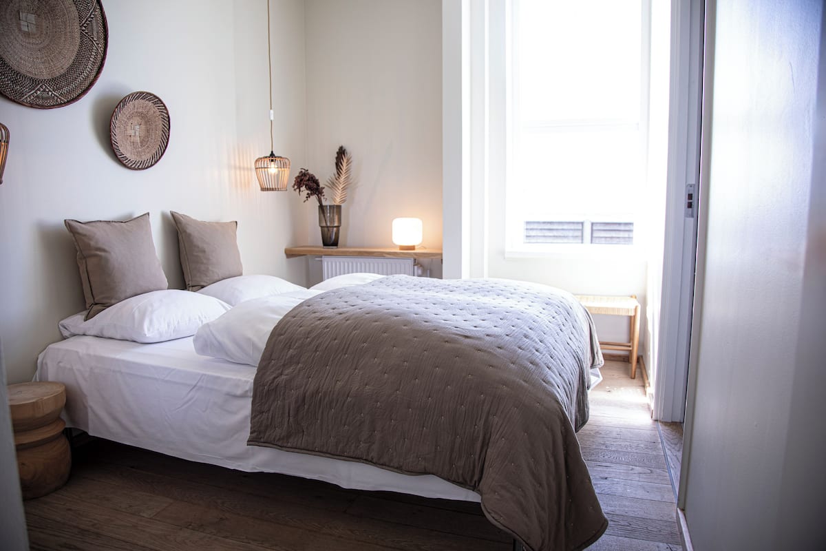 A bedroom with a bed, pillows, a bedside table with a lamp, a wall hanging, and a window.