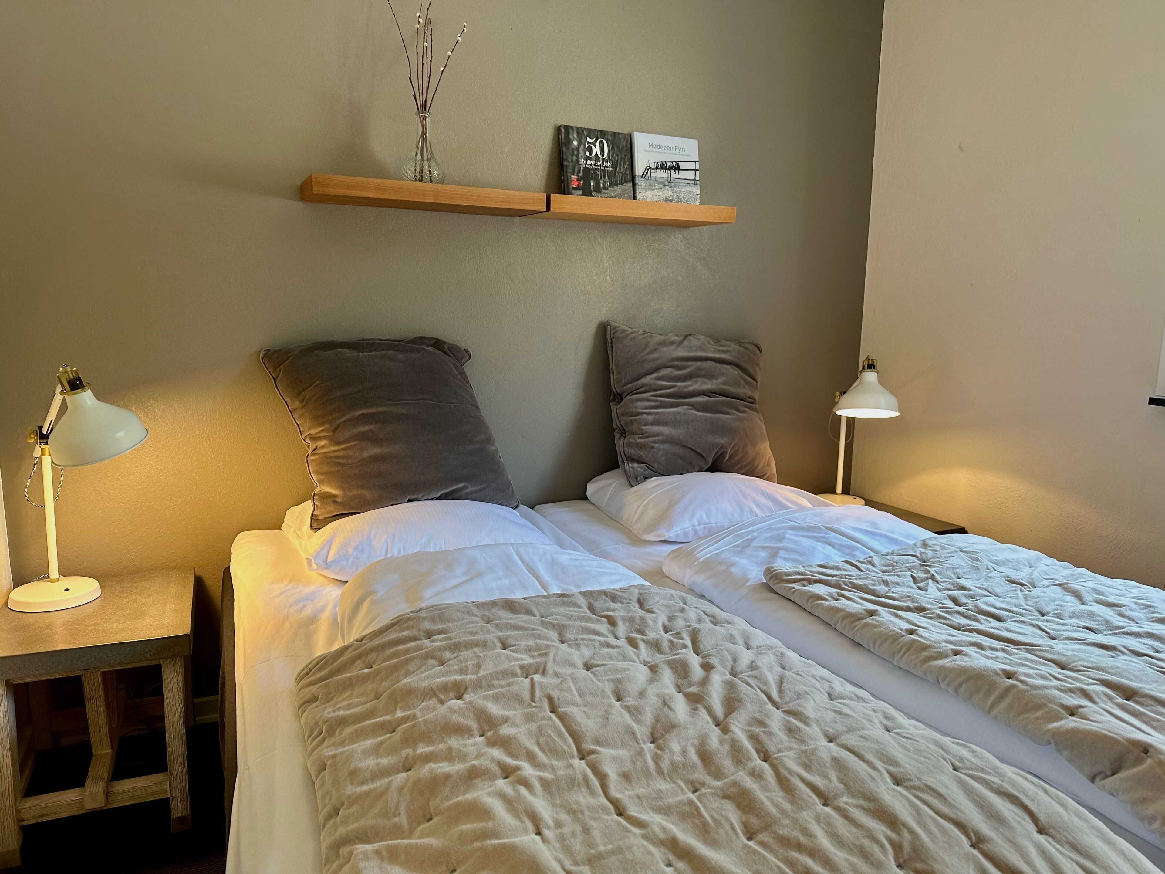 Un dormitorio con dos camas, una mesita de noche, una lámpara, un estante con libros y un florero con ramas.