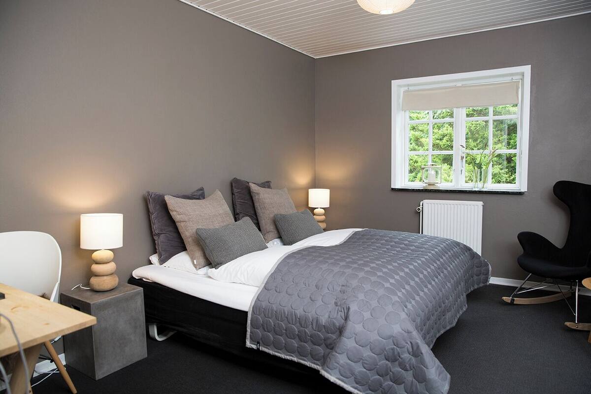 A bedroom with a bed, bedside lamps, a chair, a table, and a window with a view of greenery.