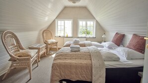 Et soveværelse med skråvæg, to vinduer, en seng med et beige dynebetræk, en fletstol og et lille bord.
