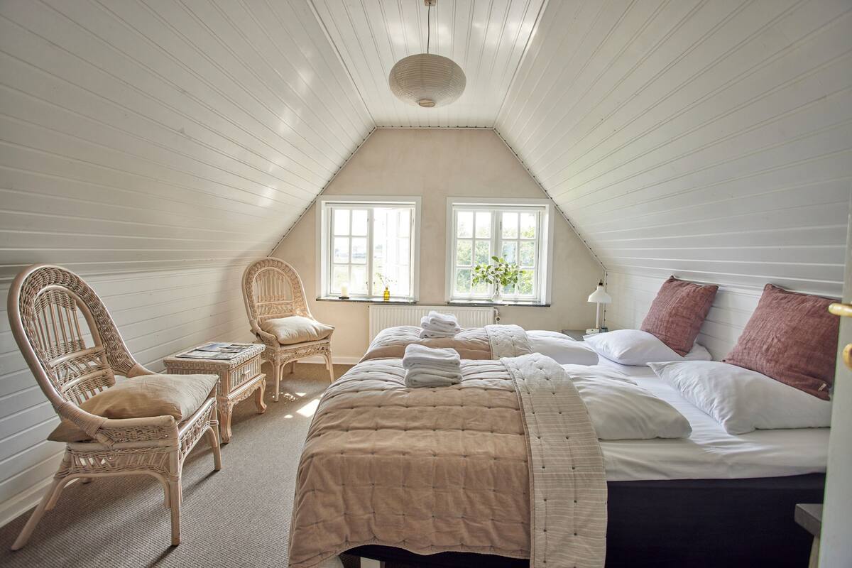 A bedroom with a sloped ceiling, two windows, a bed with a beige quilt, a wicker chair, and a small table.