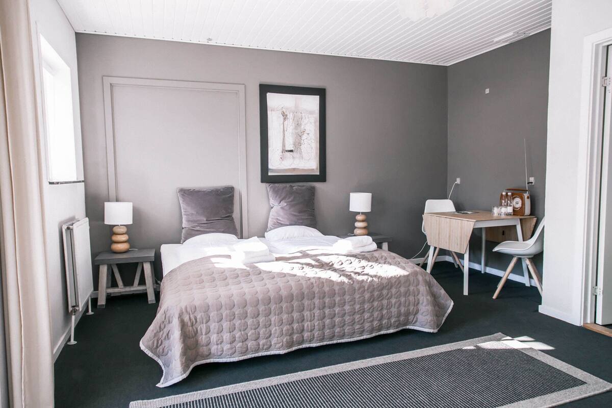 A modern bedroom with a large bed, two bedside tables, a desk with a chair, and a framed artwork on the wall.