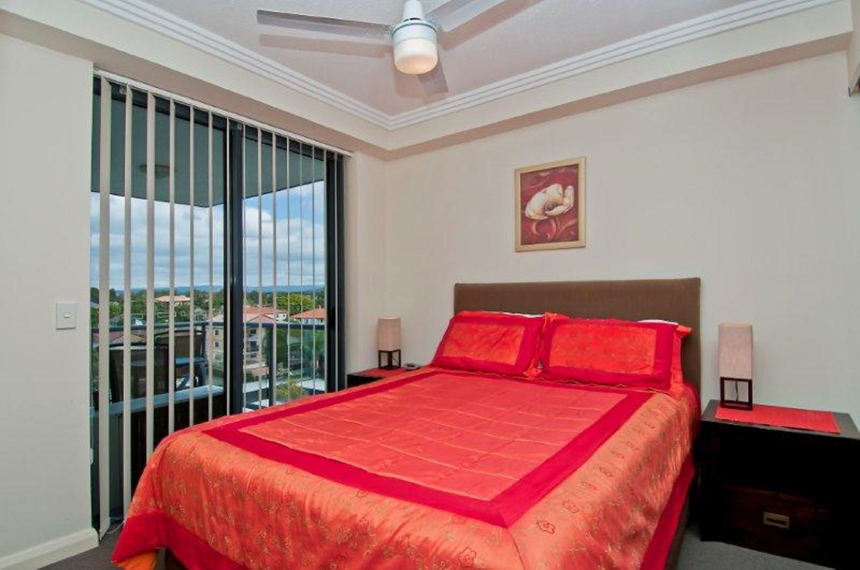 A bedroom with a large bed, a ceiling fan, a view through a sliding door, and a painting on the wall.