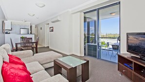 A modern living room with a sofa, a coffee table, a TV, and a balcony with outdoor furniture. - Aqualine Apartments (Southport)
