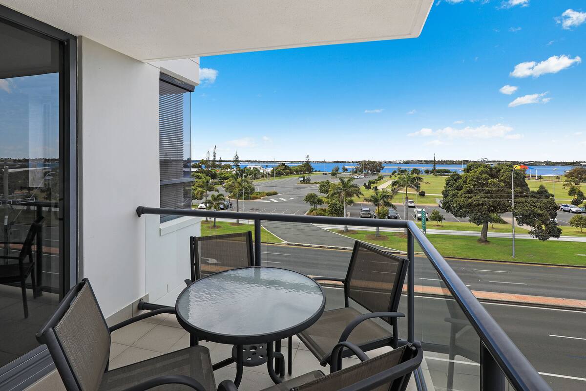 A balcony with a glass table and chairs, overlooking a road and greenery.