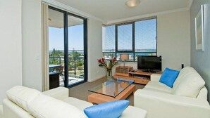 A modern living room with a glass coffee table, white sofas, a flat-screen TV, and large windows offering a view of the outdoors. - Aqualine Apartments (Southport)