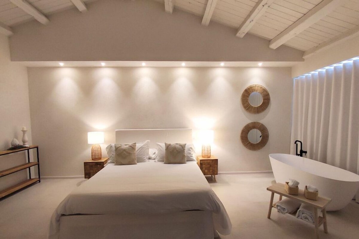 Un dormitorio con una cama grande, lámparas de noche, un espejo redondo, una bañera y un banco de madera.