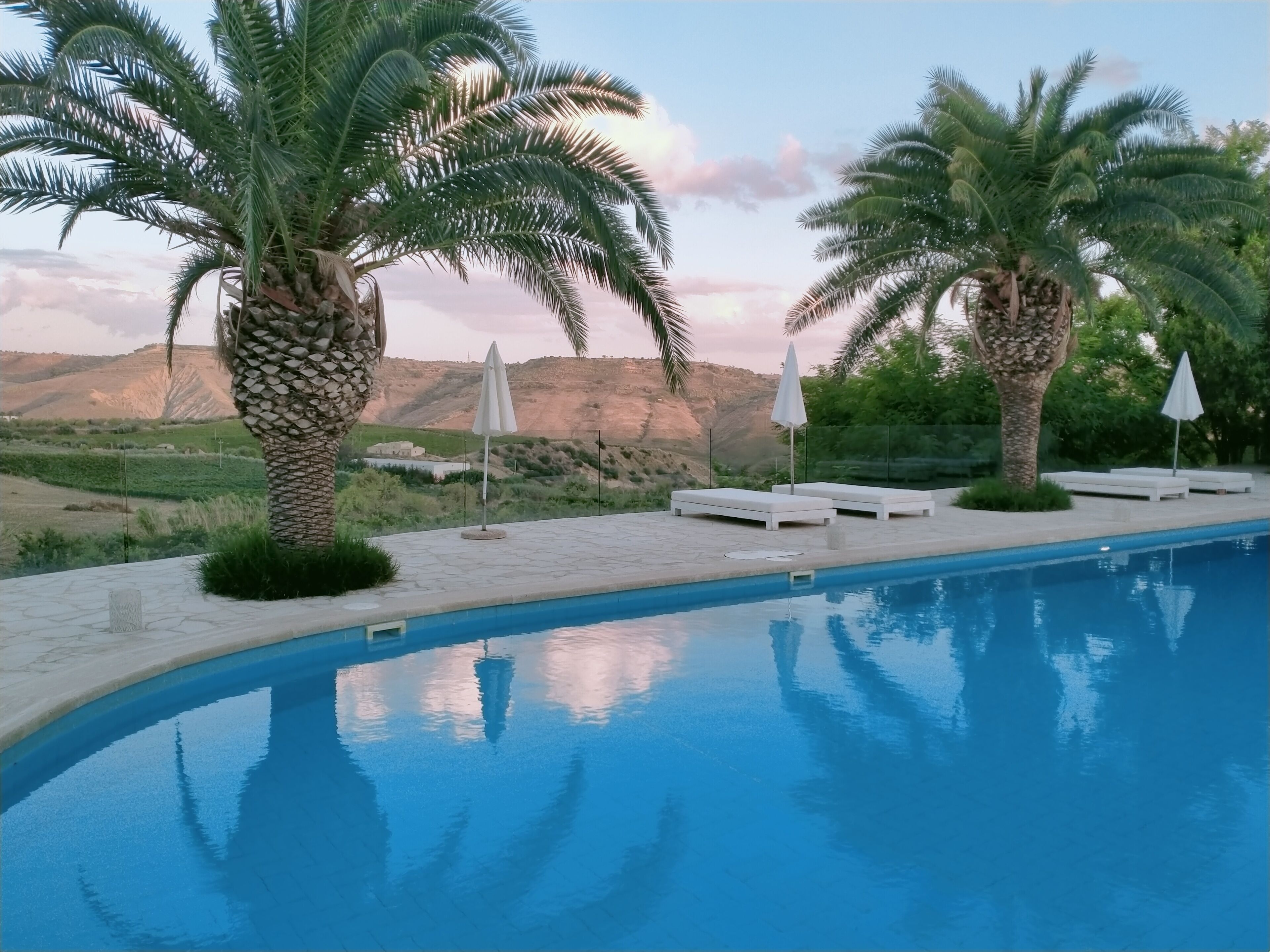 Una piscina con lettini e ombrelloni, circondata da palme e vista su un paesaggio montuoso.