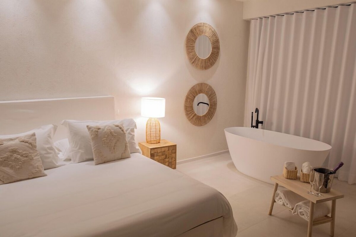 A bedroom with a large bed, a bedside lamp, a wooden nightstand, a round mirror, a bathtub, and a small table with a bottle and glasses.