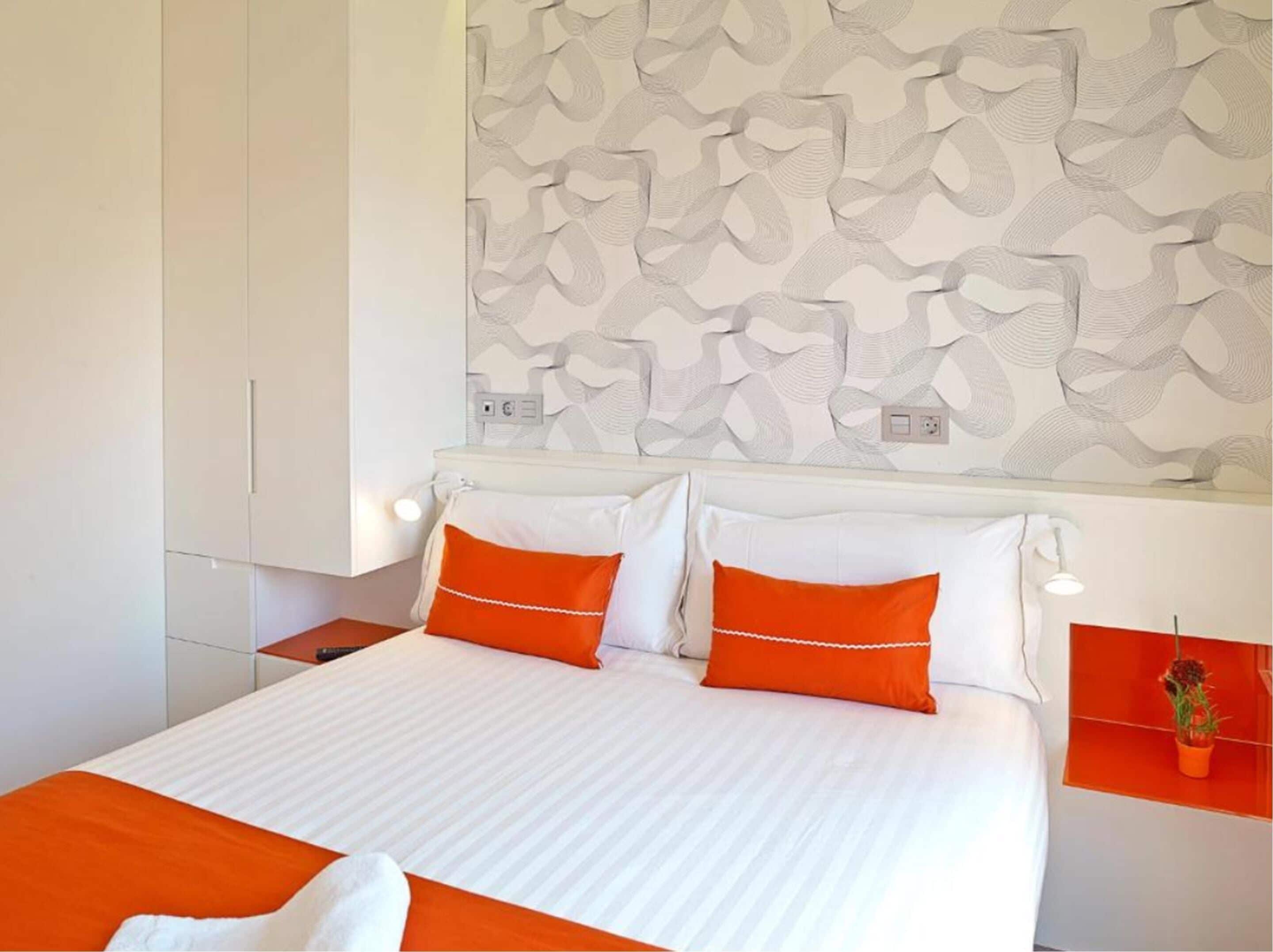 A modern hotel room with a large bed, white and orange bedding, a patterned wall, and a small plant on a shelf.