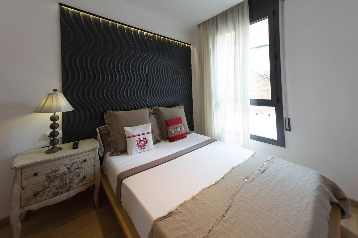 Una cama bien hecha con colcha blanca y beige, un cabecero oscuro y texturizado, y una mesita de noche con una lámpara y un objeto decorativo.