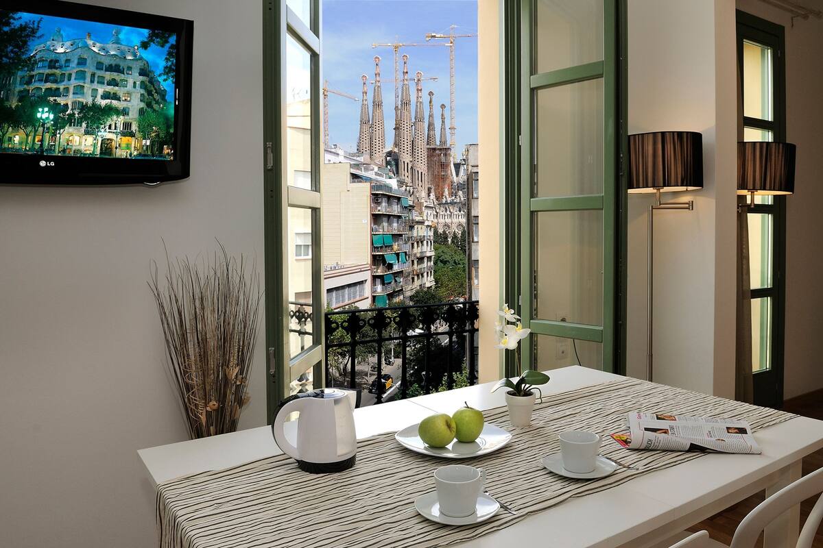 Habitación de hotel con balcón, una mesa con mantel a rayas, un plato con dos manzanas, una taza, un hervidor y una revista.
