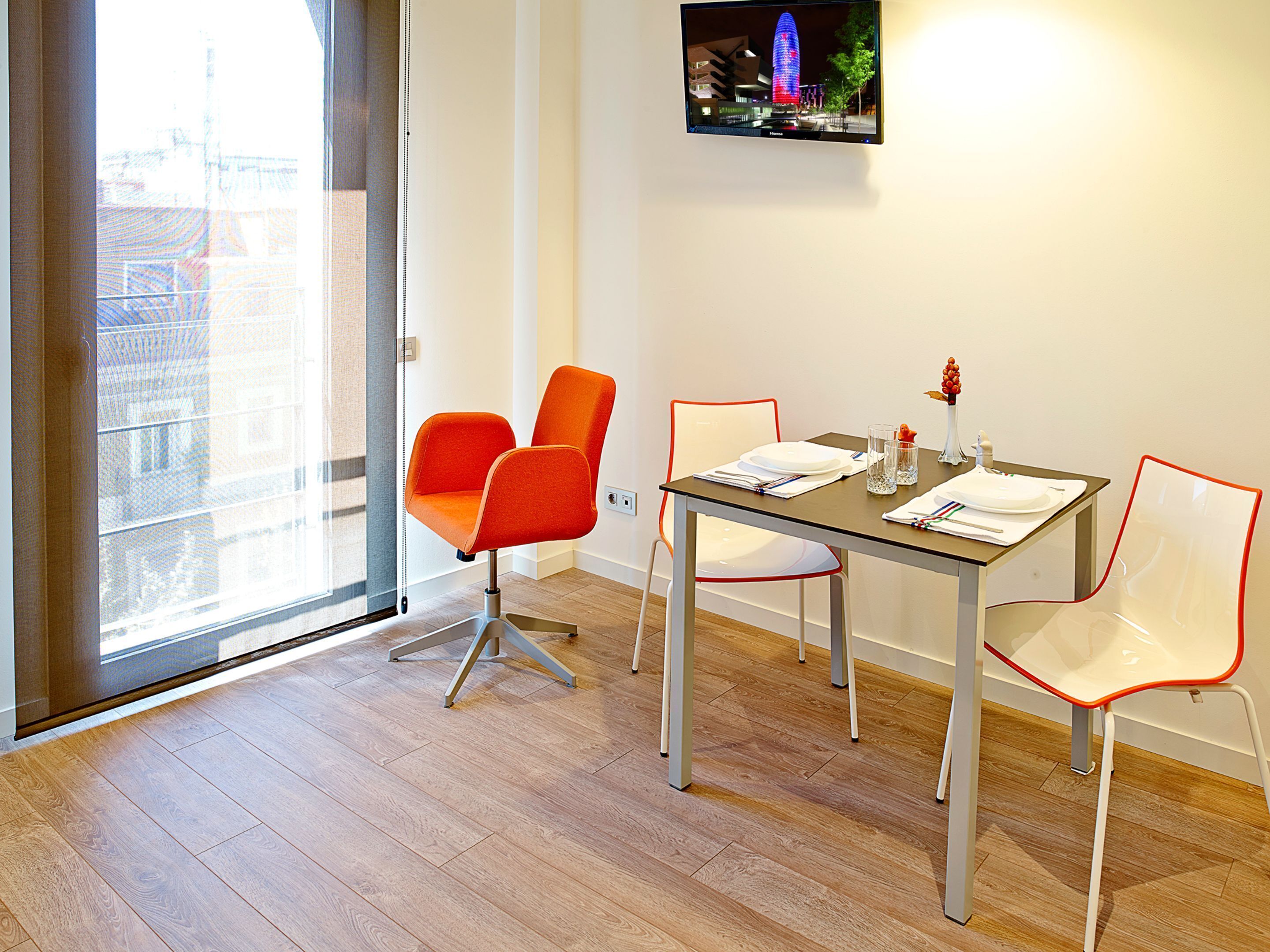 A modern room with a dining table, two orange chairs, a white chair, and a large window offering a city view.