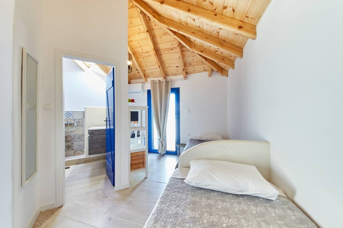 A bedroom with a wooden ceiling, a bed, and a view of a bathroom and kitchen.