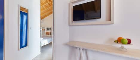 A room with a bed, a desk with a fruit bowl, a TV on the wall, and a door leading to another room.