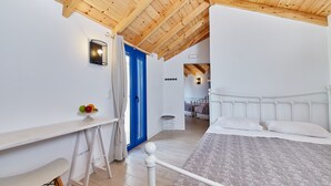 A bedroom with a wooden ceiling, a bed with white bedding, a desk with a chair, and a blue door. - Atheras (Ikaria)