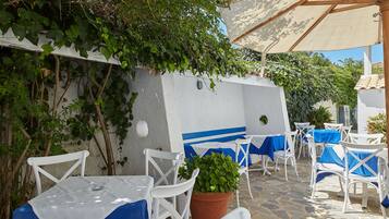 An outdoor seating area with white chairs and tables, covered by a canopy, and surrounded by greenery.