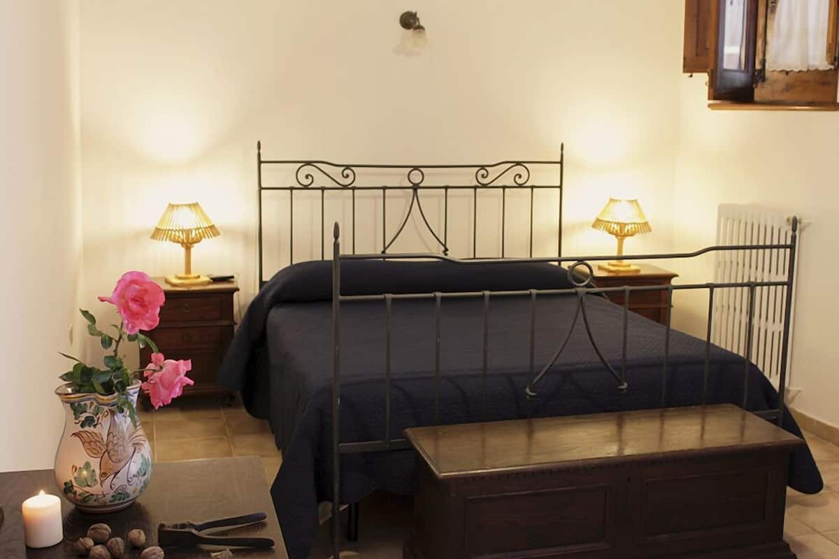 Un dormitorio con estructura de cama metálica, una mesita de noche con lámpara, un florero con flores y un radiador.