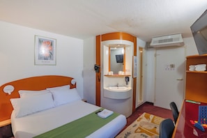 Quarto de hotel com cama, pia, espelho, porta e escrivaninha com cadeira.