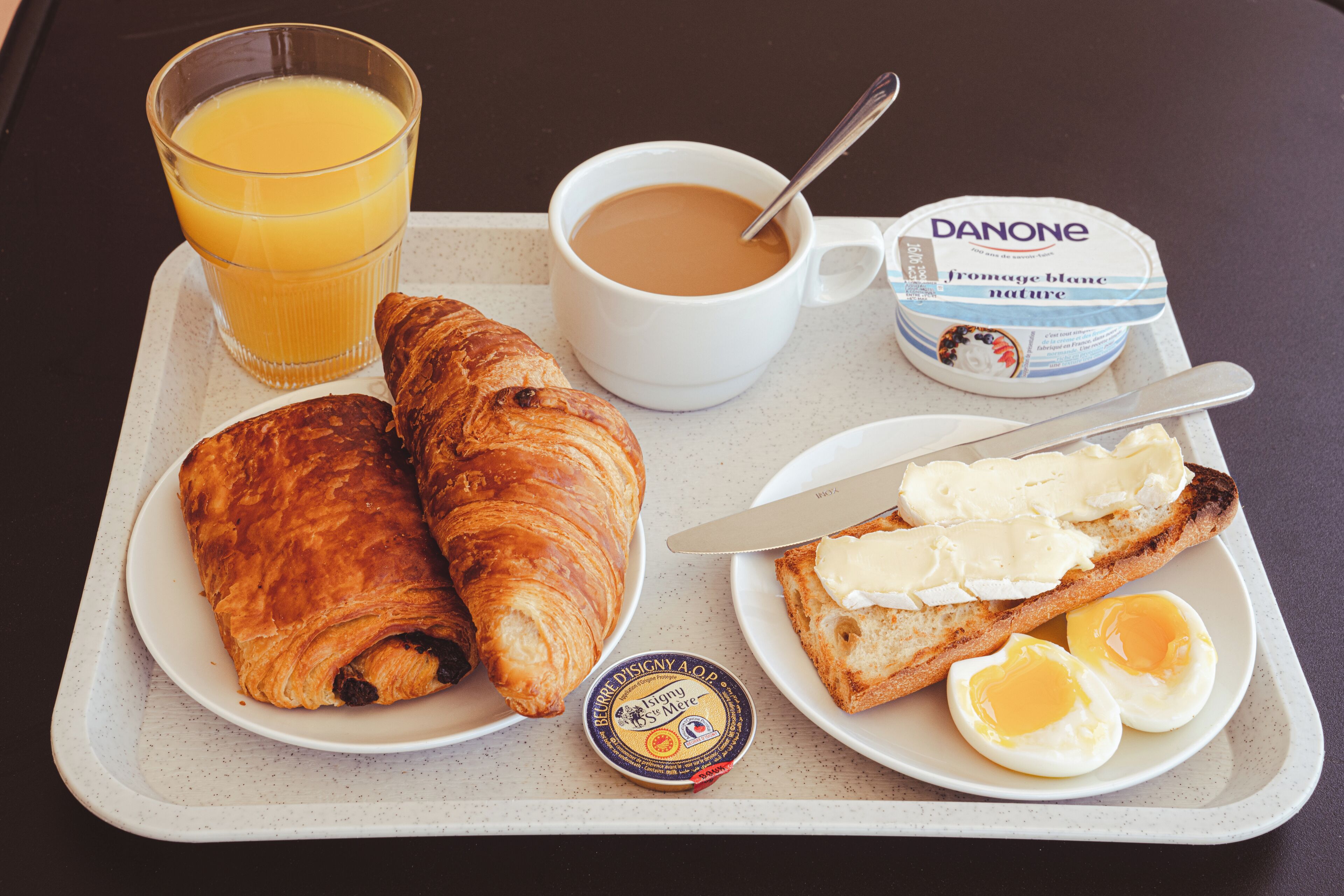 Una bandeja de desayuno con croissants, huevos, mantequilla, un vaso de zumo de naranja, una taza de café y un envase de yogur Danone.