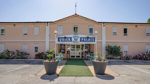 Un hotel llamado 'Quick Palace' con una entrada central, dos grandes macetas y un camino de c ésped artificial verde.