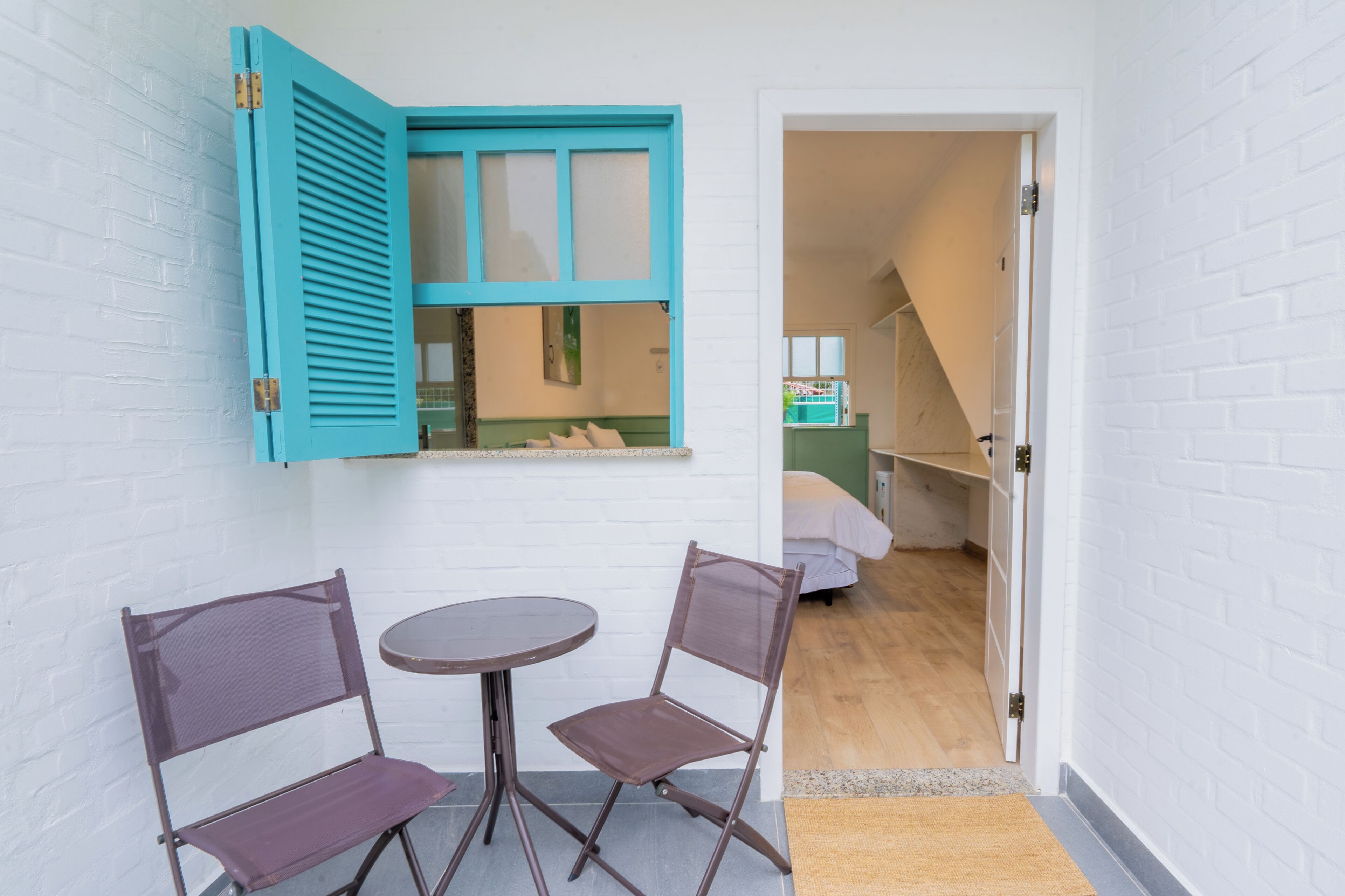 Una habitación pequeña con pared de ladrillo blanco, una ventana con contraventanas azules, una mesa redonda y dos sillas.