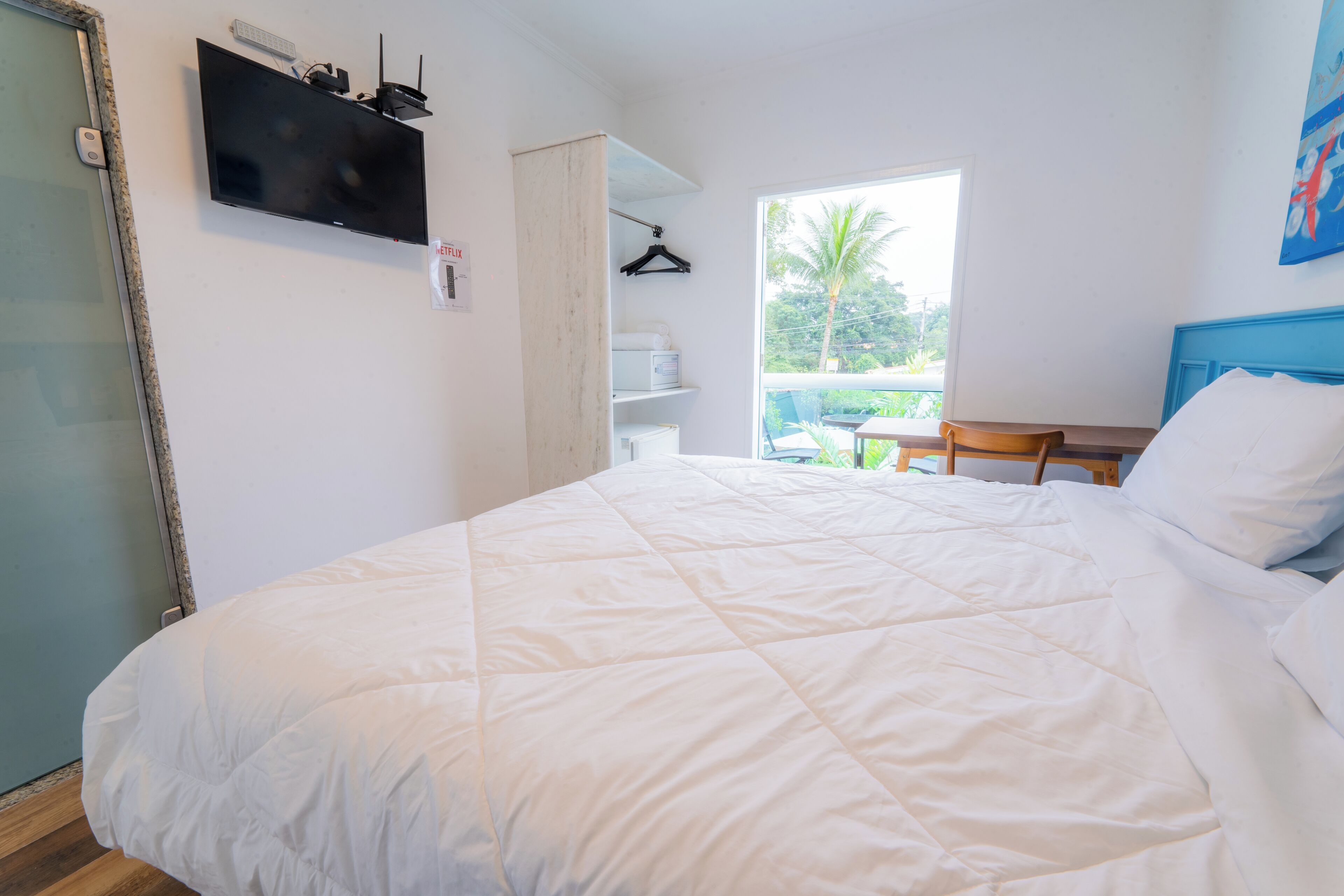 A bedroom with a large bed, a desk, and a chair. There is a window with a view of greenery outside.
