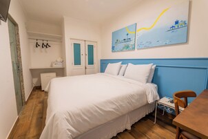 Een slaapkamer met een bed, een bureau en een stoel. Er is een raam met zonwering, een radiator en een plank met spullen erop.