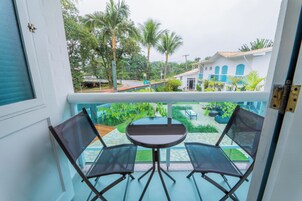 Varanda com duas cadeiras e uma mesa, com vista para um jardim com palmeiras e um edifício com janelas azuis.
