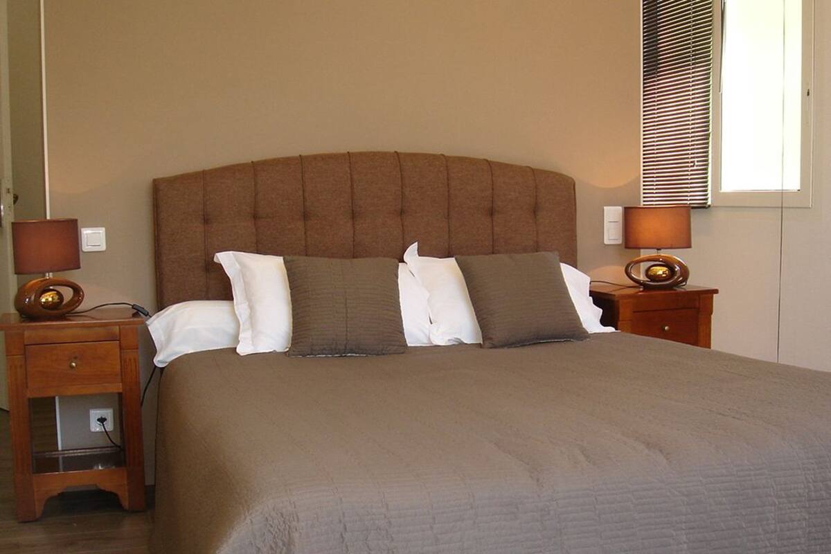 A neatly made bed with a tufted headboard, two bedside tables with lamps, and a window with blinds.