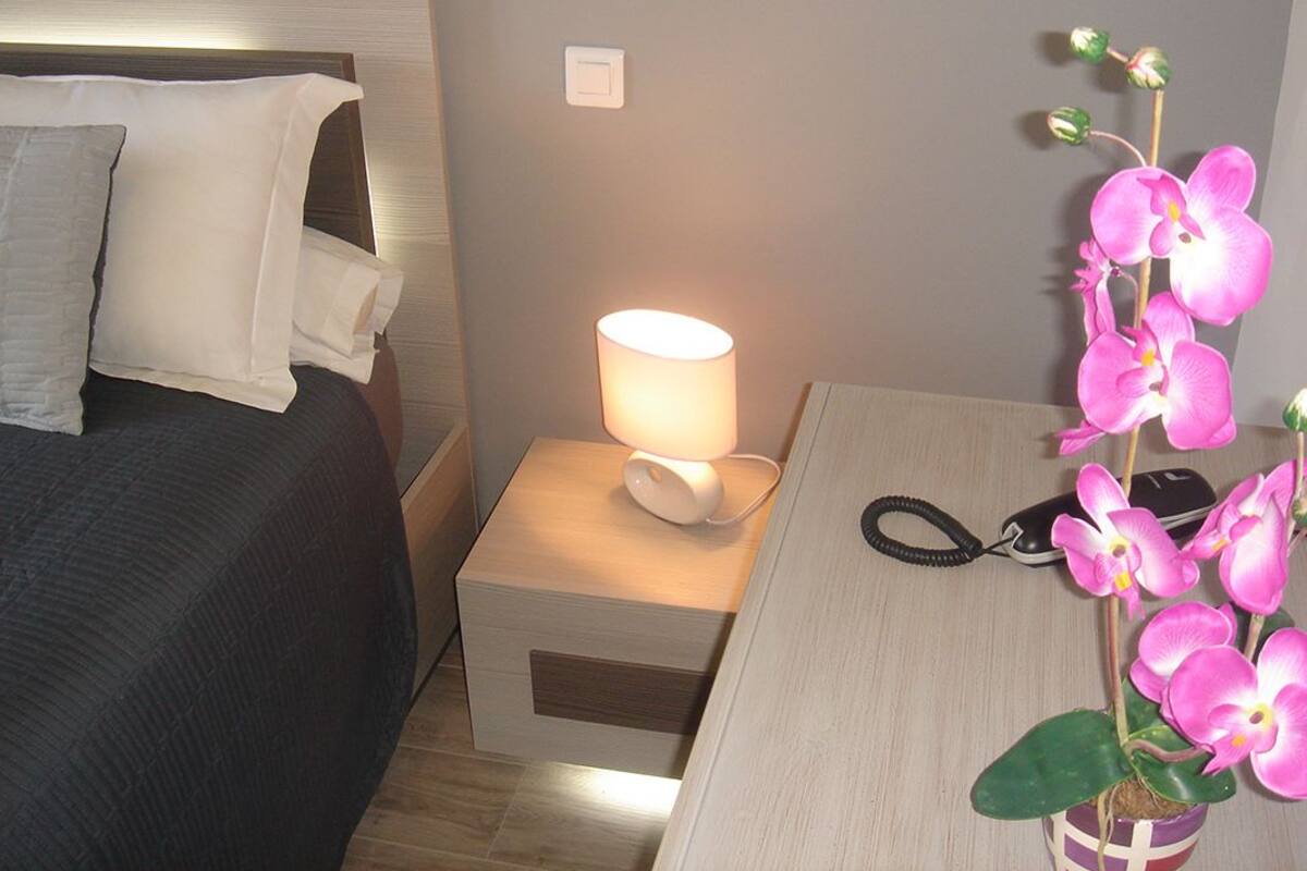 A neatly made bed with white linens, a bedside lamp, a small table with a phone and a potted orchid, and a nightstand with a drawer.