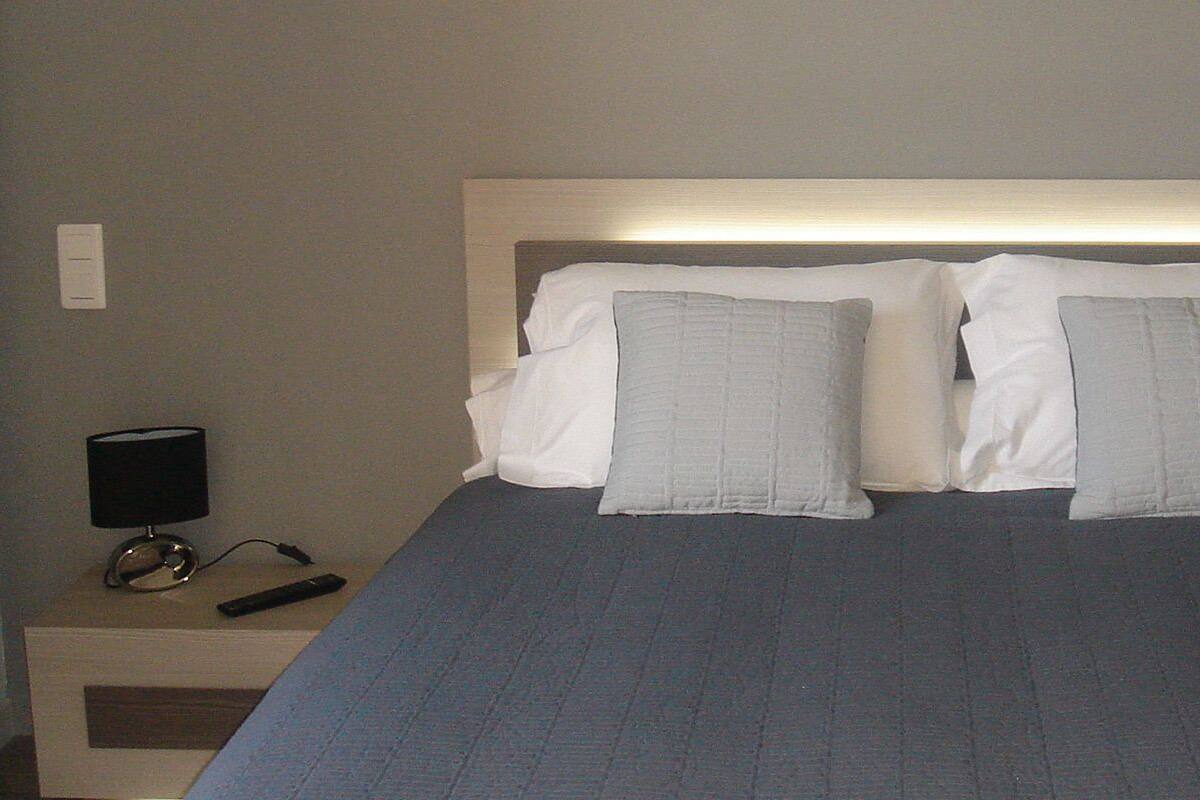 A neatly made bed with a grey bedspread and two pillows, a bedside lamp, and a remote control on a wooden nightstand.