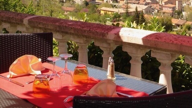 A table with a red tablecloth, a bottle, and glasses, overlooking a town with mountains in the background. - Auberge Provencale (Sospel)